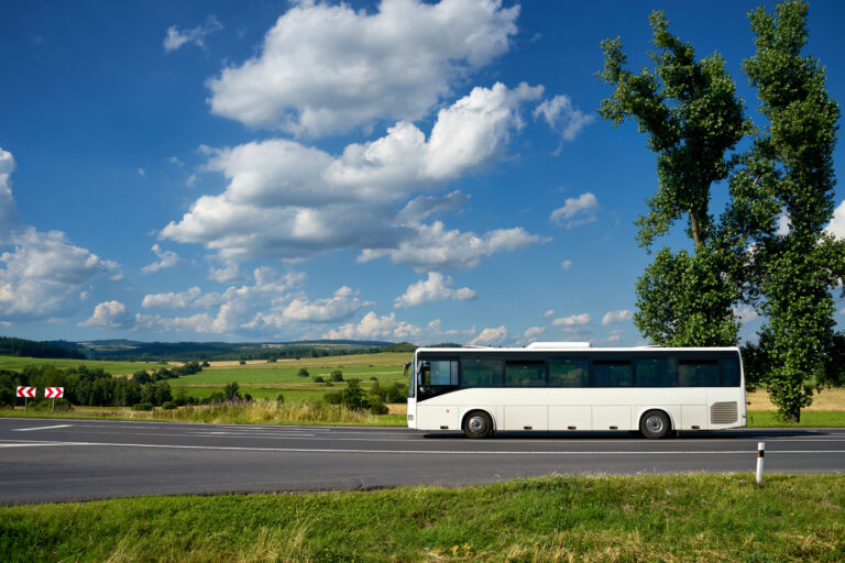 Ein weißer Bus fährt über eine Straße vor grüner, sonniger Landschaft